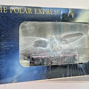 May include: A Lionel The Polar Express elf handcar model train set in a rectangular cardboard box. The packaging features a dark blue snowy night scene, the text The Polar Express, and a clear plastic window displaying the maroon handcar inside.