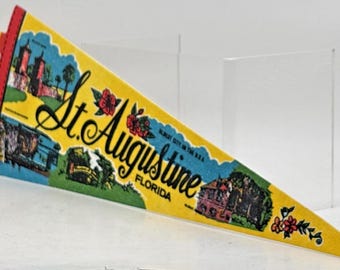 May include: A triangular felt pennant featuring a yellow background with the text St. Augustine Florida in black script. The design includes colorful illustrations of historic buildings, palm trees, and red flowers, with two yellow fabric ties at the base.