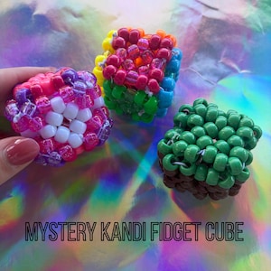 Puede incluir: Tres coloridos cubos fidget Kandi. Un cubo es rosa, morado y blanco. Otro es de color arco&iacute;ris, y el tercero es verde y marr&oacute;n. El texto "MYSTERY KANDI FIDGET CUBE" est&aacute; en la parte inferior.