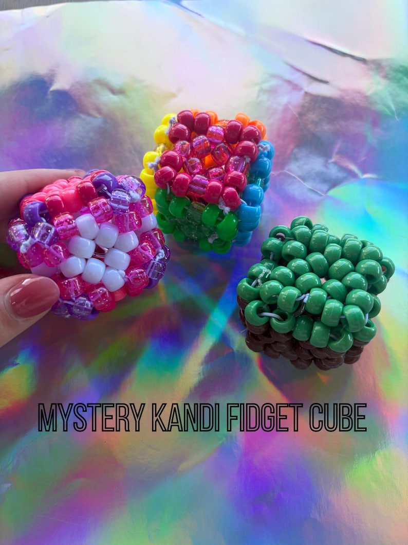 Puede incluir: Tres coloridos cubos fidget Kandi. Un cubo es rosa, morado y blanco. Otro es de color arco&iacute;ris, y el tercero es verde y marr&oacute;n. El texto "MYSTERY KANDI FIDGET CUBE" est&aacute; en la parte inferior.