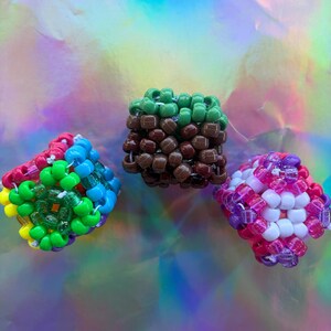 Puede incluir: Tres creaciones c&uacute;bicas de cuentas de colores. Una es una mezcla de cuentas rojas, verdes, azules y amarillas. Otra es verde y marr&oacute;n, y la tercera es rosa, morada y blanca. El fondo es una superficie hologr&aacute;fica iridiscente.