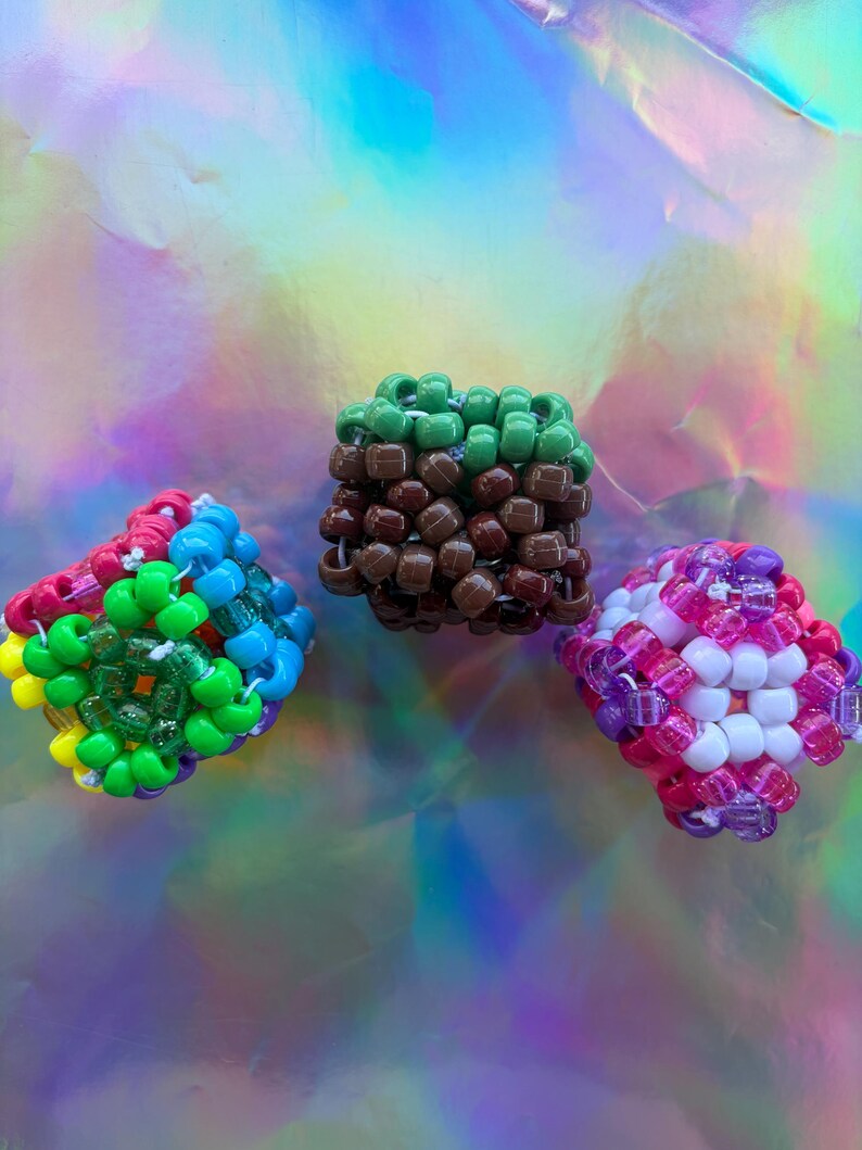 Puede incluir: Tres creaciones c&uacute;bicas de cuentas de colores. Una es una mezcla de cuentas rojas, verdes, azules y amarillas. Otra es verde y marr&oacute;n, y la tercera es rosa, morada y blanca. El fondo es una superficie hologr&aacute;fica iridiscente.