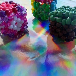 Puede incluir: Tres pulseras de cuentas de colores. Una pulsera presenta cuentas rosas, moradas y blancas. Otra tiene un arco&iacute;ris de colores, incluyendo amarillo, verde y azul. La tercera pulsera es marr&oacute;n y verde. Las pulseras est&aacute;n sobre una superficie reflectante e iridiscente.