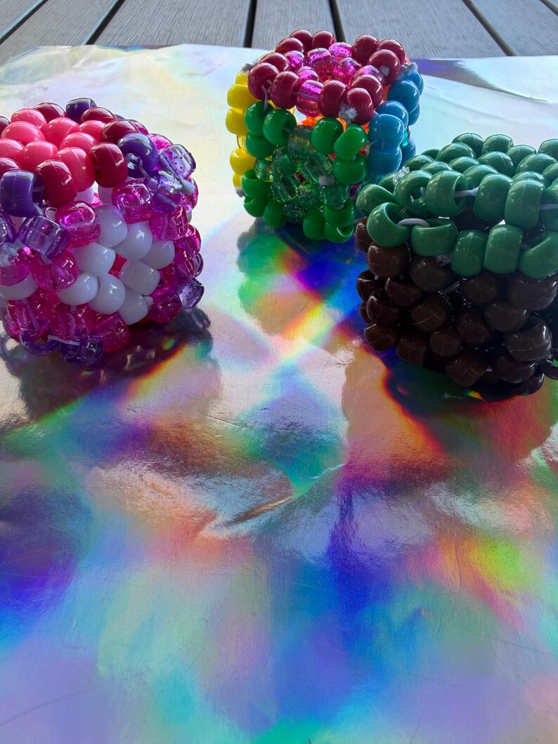 Puede incluir: Tres pulseras de cuentas de colores. Una pulsera presenta cuentas rosas, moradas y blancas. Otra tiene un arco&iacute;ris de colores, incluyendo amarillo, verde y azul. La tercera pulsera es marr&oacute;n y verde. Las pulseras est&aacute;n sobre una superficie reflectante e iridiscente.