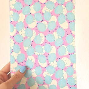 Könnte beinhalten: Ein Notizbuch mit rosa Hintergrund und einem Muster aus hellblauen Formen und cremefarbenen Ovalen. Das Notizbuch wird in einer Hand mit hellblauem Nagellack gehalten.