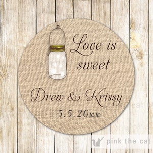 May include: A round, burlap-textured wedding favor tag with the text "Love is sweet" and "Drew & Krissy" with the date "5.5.20xx". A small mason jar with a lit candle inside is pictured above the text.