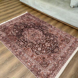 May include: A rectangular, ornate rug with a central floral medallion design in shades of brown, burgundy, and cream. The rug features a detailed border and a white fringe. The rug is placed on a wooden floor.