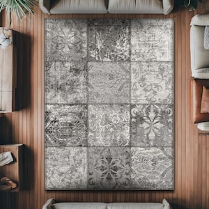 Puede incluir: Una alfombra rectangular grande con un diseño de patchwork. La alfombra presenta una variedad de patrones grises y blancos, incluyendo motivos florales y abstractos. La alfombra está colocada sobre un suelo de madera, rodeada de muebles.