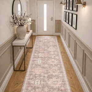 May include: A long, rectangular rug with a faded, patterned design in shades of mauve, beige, and orange. The rug is placed in a hallway with hardwood flooring and white walls. The hallway also features a white console table and framed artwork.