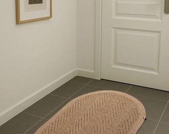May include: Oval-shaped, beige rug with a woven chevron pattern. The rug is placed on a tiled floor near a white door and a framed abstract art piece. The rug adds a touch of warmth and texture to the space.