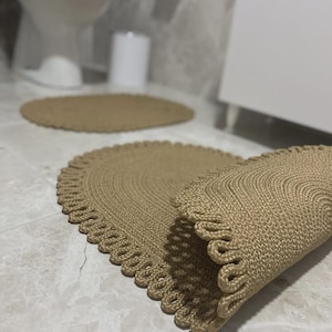 Könnte beinhalten: Ein Set aus drei beigefarbenen, gewebten Badezimmerteppichen. Zwei sind oval geformt mit einem gewellten Rand, und einer ist aufgerollt. Die Teppiche bestehen aus einem strukturierten Material und liegen auf einem gefliesten Boden.