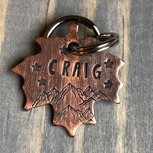 May include: Copper leaf-shaped dog tag with the name "CRAIG" engraved in black lettering. The tag features a mountain range design and five small stars.