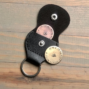 May include: Black leather keychain with two metal golf ball markers. One marker is copper-coloured with the text "FOREVER TO GO" and the date "6.15.18". The other marker is gold-coloured with the text "FOREVER TO GO" and the date "5.19.17".