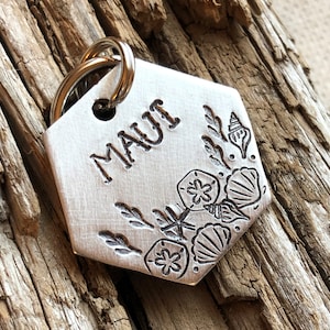 May include: Silver hexagonal pet tag with the word "MAUI" engraved on it. The tag is decorated with a seashell and starfish design.