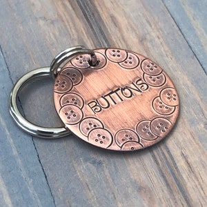 May include: A copper dog tag with a circle of buttons around the edge and the word "BUTTONS" engraved in the centre. The tag is attached to a silver key ring.