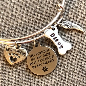 Op de afbeelding: Zilveren armband met een hartvormige bedel met de tekst "Beste vriend" met poot afdrukken, een bedel in de vorm van een bot met de tekst "Bitsy", een vleugelbedel en een ronde bedel met de tekst "Niet langer aan mijn zijde, maar voor altijd in mijn hart" met een poot afdruk.