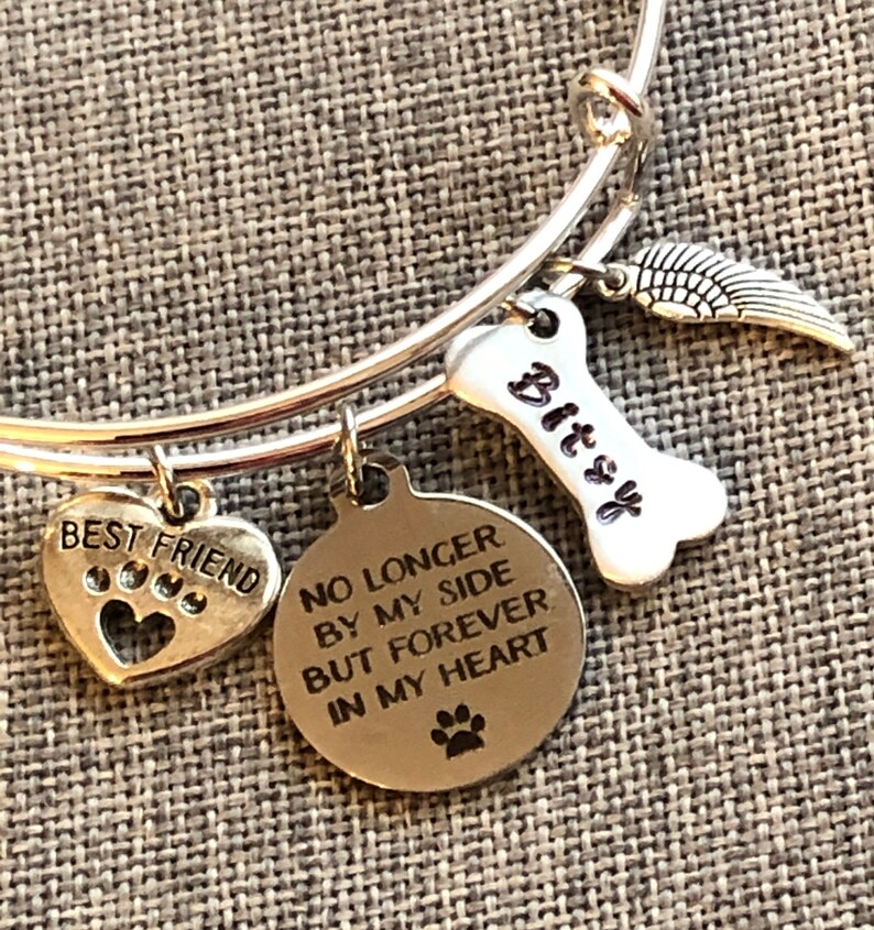 Op de afbeelding: Zilveren armband met een hartvormige bedel met de tekst "Beste vriend" met poot afdrukken, een bedel in de vorm van een bot met de tekst "Bitsy", een vleugelbedel en een ronde bedel met de tekst "Niet langer aan mijn zijde, maar voor altijd in mijn hart" met een poot afdruk.