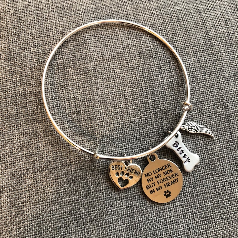 Op de afbeelding: Zilveren armband met een hartvormige bedel met een pootprint, een bedel in de vorm van een bot met de naam "Bitsy" erop gegraveerd en een bedel met de tekst "Niet langer aan mijn zijde, maar voor altijd in mijn hart".