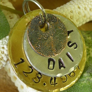 May include: A layered metal pendant with a silver ring. The pendant features three circular layers: a gold-toned base with stamped numbers, a silver middle layer with the name "DAIS" stamped, and a copper top layer with a floral design.