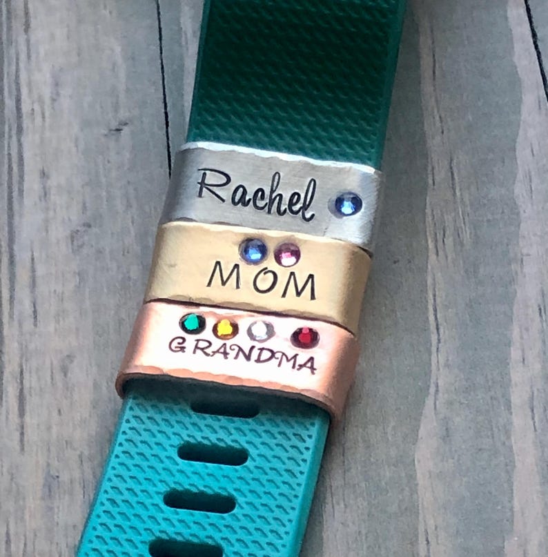 May include: Three metal charms with rhinestones, engraved with the words "Rachel", "MOM", and "GRANDMA". The charms are attached to a teal fitness tracker band.