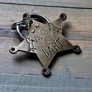 May include: A brass star-shaped dog tag with a cowboy theme. The tag features a cactus, a horse, and a cowboy. The name "BART" is engraved on the tag.