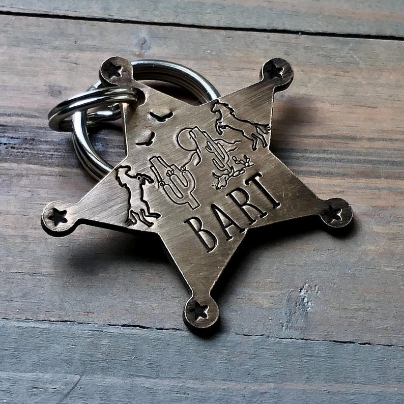 May include: A brass star-shaped dog tag with a cowboy theme. The tag features a cactus, a horse, and a cowboy. The name "BART" is engraved on the tag.