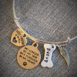 Op de afbeelding: Zilveren armband met een hartvormige bedel met de tekst "Beste vriend" en een ronde bedel met de tekst "Niet langer aan mijn zijde, maar voor altijd in mijn hart". Er is ook een bedel in de vorm van een bot met de tekst "Bitsy" en een kleine engelenvleugelbedel.
