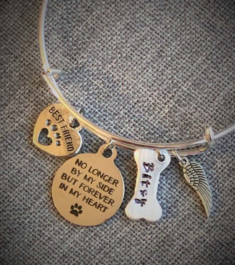 Op de afbeelding: Zilveren armband met een hartvormige bedel met de tekst "Beste vriend" en een ronde bedel met de tekst "Niet langer aan mijn zijde, maar voor altijd in mijn hart". Er is ook een bedel in de vorm van een bot met de tekst "Bitsy" en een kleine engelenvleugelbedel.