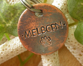 Może przedstawiać: Miedziana plakietka dla psa z wygrawerowanym imieniem "MELBORN." Plakietka ma postarzony wygląd i wzór odcisku łapy.