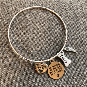 Op de afbeelding: Zilveren armband met een hartbedel met de tekst "Beste vriend" met een pootprint, een ronde bedel met de tekst "Niet langer aan mijn zijde, maar voor altijd in mijn hart" met een pootprint, een bedel in de vorm van een bot met de tekst "Bitzy" en een vleugelbedel.