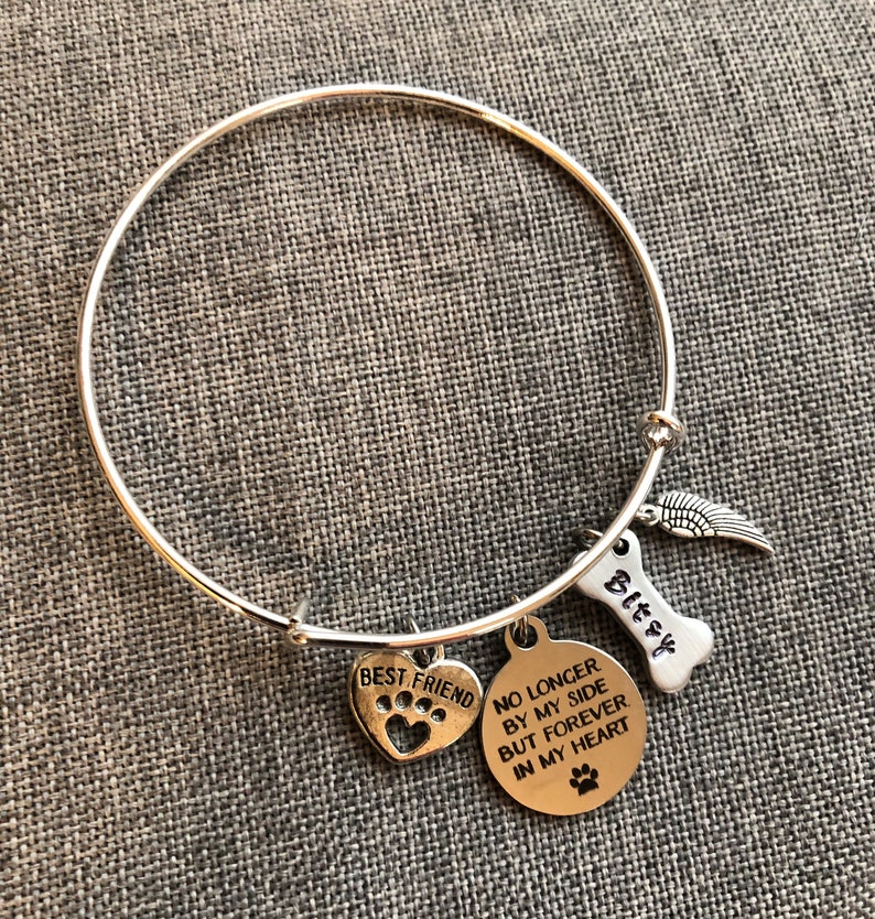 Op de afbeelding: Zilveren armband met een hartbedel met de tekst "Beste vriend" met een pootprint, een ronde bedel met de tekst "Niet langer aan mijn zijde, maar voor altijd in mijn hart" met een pootprint, een bedel in de vorm van een bot met de tekst "Bitzy" en een vleugelbedel.