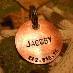 May include: A copper dog tag with the name "JACOBY" and the phone number "212.518.16" engraved on it.