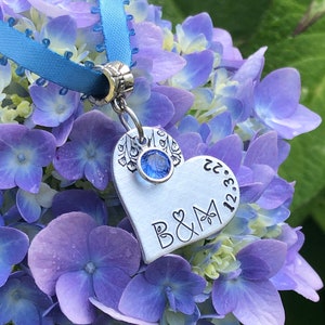 May include: A silver heart charm with a blue crystal and the inscription "B&M 12.23.22" hanging from a blue ribbon against a backdrop of purple hydrangeas.