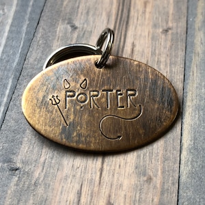 May include: Oval-shaped, bronze-colored pet tag with the name "PORTER" engraved in the center. A trident and a devil's tail are etched on either side of the name. The tag is attached to a split ring for easy attachment to a pet's collar.