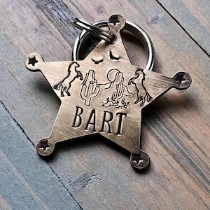 May include: A gold-toned metal sheriff's star keychain with a cowboy theme. The star has a cutout design of two horses, a cactus, and a mountain. The name "BART" is engraved on the star.