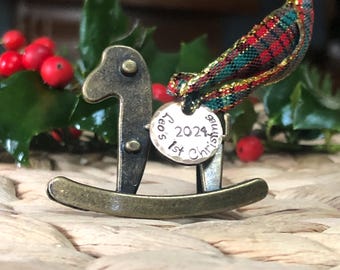 May include: A small, gold-colored rocking horse ornament with a silver tag that reads "2024,  Féo's 1st Christmas". The ornament is surrounded by green holly with red berries.