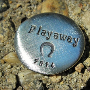 May include: A silver metal token with the words "Playaway" and the year "2014" engraved on it. The token also features a stylized omega symbol.