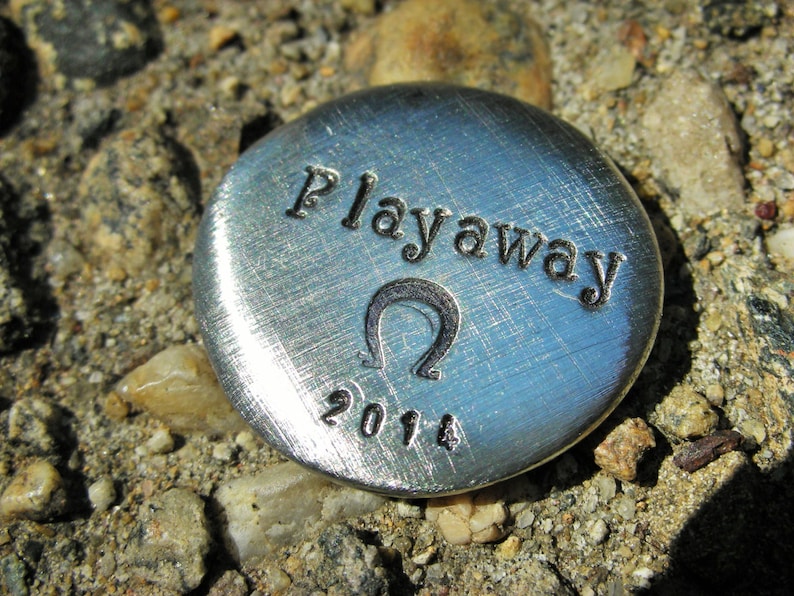 May include: A silver metal token with the words "Playaway" and the year "2014" engraved on it. The token also features a stylized omega symbol.
