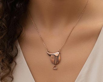 May include: A copper-colored necklace featuring a pendant shaped like a horse saddle. The pendant has a stirrup and a curved top. The chain is delicate and appears to be the same color as the pendant. The necklace is worn against a light background.