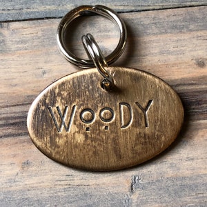 May include: A brass oval dog tag with a silver key ring. The tag has the name "WOODY" engraved on it.