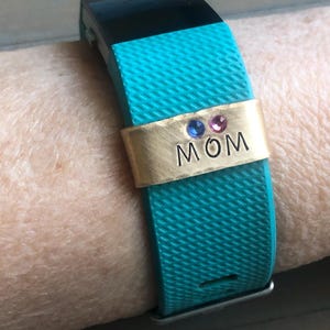 May include: A turquoise fitness tracker with a gold band that says "MOM" with three small gemstones.