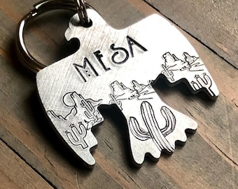 May include: A silver-colored metal keychain in the shape of a bird, with the word "MESA" engraved at the top. The bird design features desert landscape imagery, including cacti and mountains. The keychain is attached to a silver key ring.