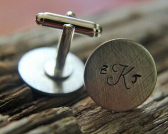 May include: A pair of silver cufflinks with a brushed finish. The cufflinks feature a round, engraved design with the initials "ZK" in a cursive font.