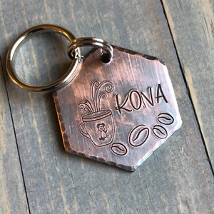 May include: A copper-colored hexagonal dog tag with a key ring. The tag has a coffee cup with steam coming out of it and a small dog inside the cup. The tag also has the word "KONA" and three coffee beans engraved on it.