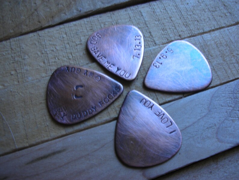May include: Four copper guitar picks with different engraved messages. The messages are "God Save Me You", "7.13.18", "I Love You", and "C.M.X. 604 Buddy Rocks".