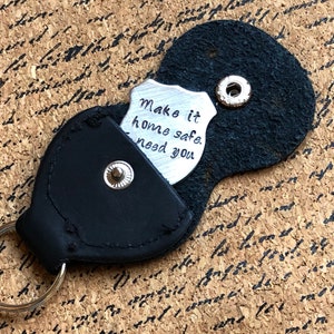 Puede incluir: Llavero de cuero negro con un dije de metal plateado en forma de escudo que dice "Make it home safe. Need you."
