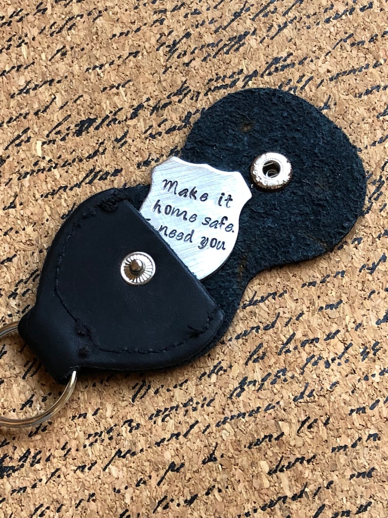 Puede incluir: Llavero de cuero negro con un dije de metal plateado en forma de escudo que dice "Make it home safe. Need you."
