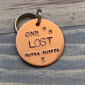 May include: Copper dog tag with a silver key ring. The tag reads "One Lost Mutha Pupper" with two paw prints and a small dog illustration.