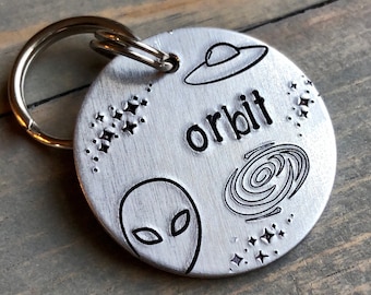 May include: Silver-toned metal dog tag with a black alien, UFO, galaxy, and stars design. The tag reads "orbit".