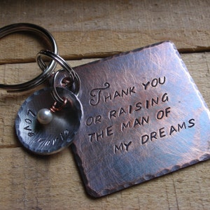 May include: A copper-toned keychain with a square tag that reads "Thank you for raising the man of my dreams". The tag is attached to a silver key ring with a small silver charm that has a pearl and the word "Love" engraved on it.